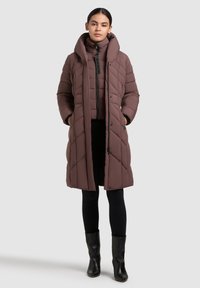 Nő hosszú mályva színű, steppelt kabátban, magas gallérral, fekete leggingsben és fekete bokacsizmában, kezét a zsebében tartva áll.