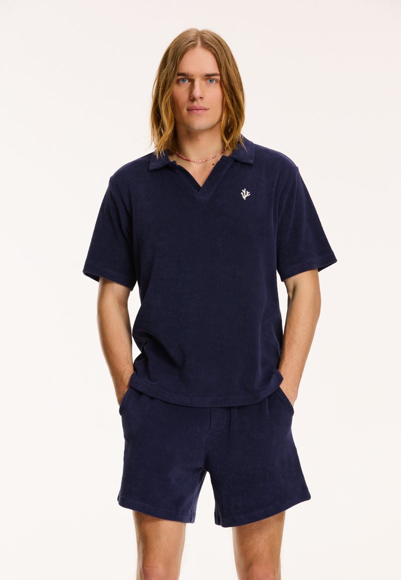 Polo shirt din terrycloth de marin, cu guler în V și mâneci scurte, având un mic emblemă în formă de frunză. Asortat cu pantaloni scurți navy.