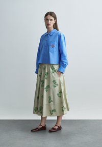 Femeie purtând o cămașă albastră cu nasturi și buzunar la piept și o fustă bej cu imprimeuri florale verzi, stând în picioare în fața unui fundal simplu.