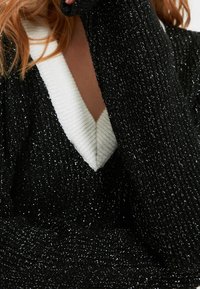 Pull en maille métallique noire et argentée avec un col en V profond et un large col blanc ; manches longues à texture côtelée.