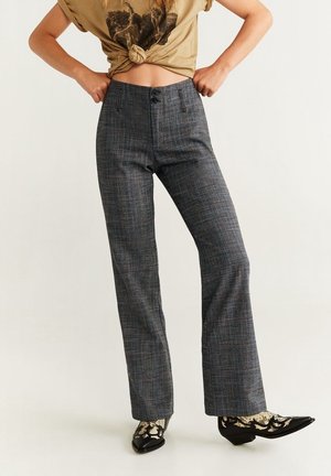 Femme portant un pantalon à carreaux gris taille haute, un t-shirt graphique noué beige, et des chaussures noires pointues avec des chaussettes beiges à motifs, debout les mains sur les hanches.