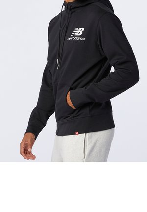 Mężczyzna ubrany w czarną bluzę New Balance na zamek, z lewą ręką w kieszeni, w jasnoszarych spodniach, na jednolitym tle.