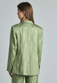 Blazer in satin verde chiaro con un motivo ondulato e texture. Presenta un rever a punta e tre bottoni su ciascun polsino, vestibilità aderente.