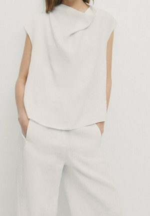Femme portant un top blanc sans manches avec col drapé et pantalon large assorti, mains dans les poches, debout sur fond blanc.