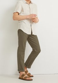 Camisa de lino de manga corta en color beige claro, chinos verde olivo y sandalias marrones de dos tiras. La camisa tiene un cuello sutil y un bolsillo en la parte delantera.