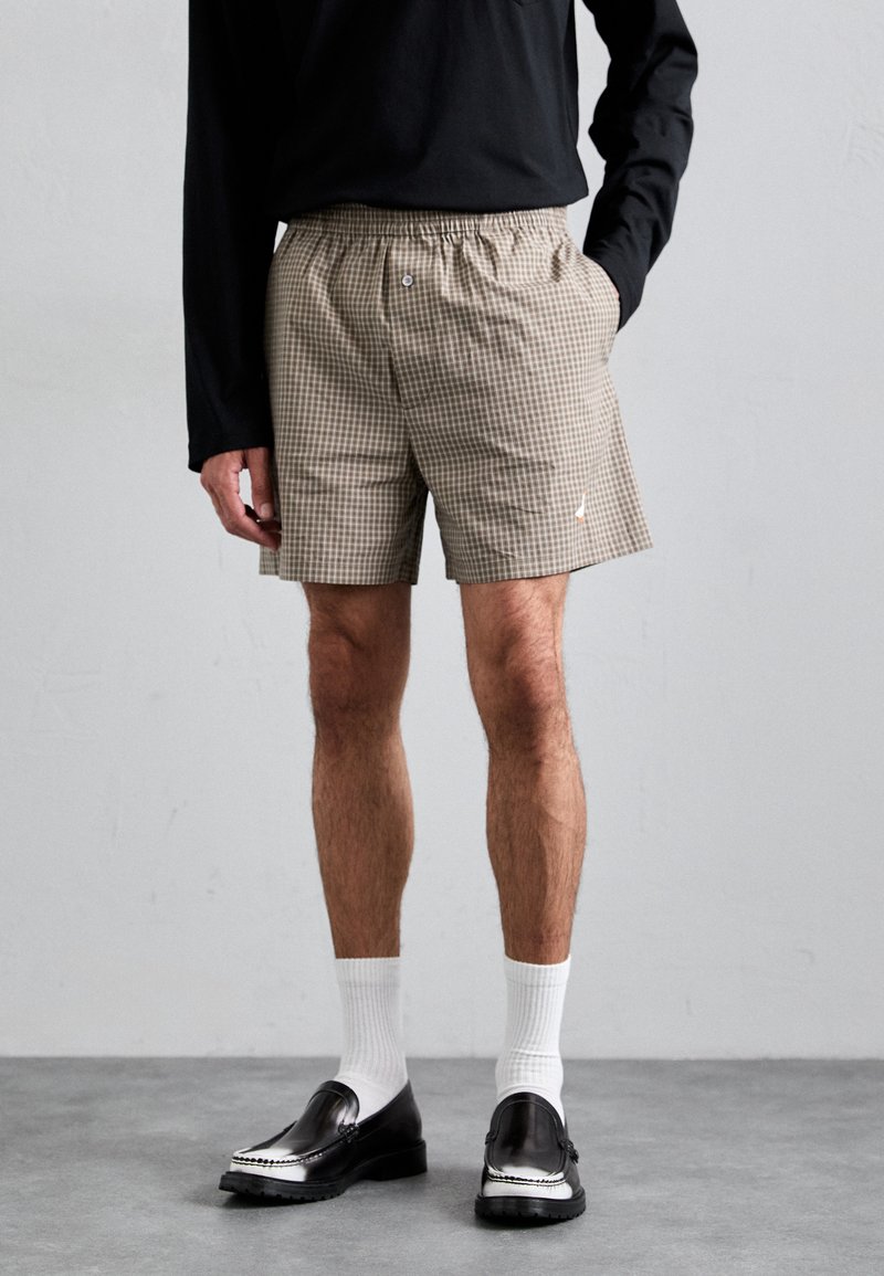 Bruine geblokte shorts met een elastische tailleband, zijkanten zakken en een klein logo-detail. Draagt met zwarte instappers en witte sokken.