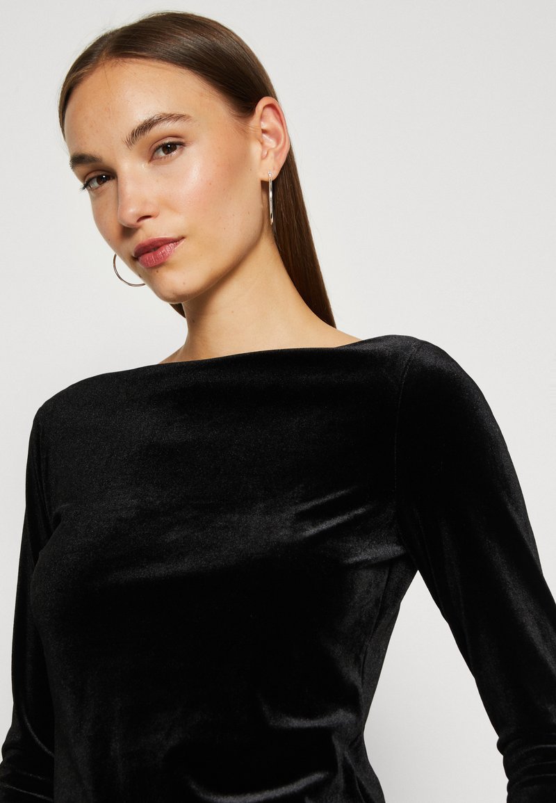 Eine Frau mit glattem braunem Haar trägt silberne Creolen und ein schwarzes, langärmliges Oberteil aus Samt. Sie posiert vor einem schlichten, hellen Hintergrund.