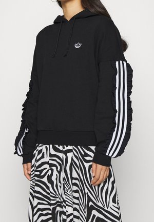 Femme portant un sweat à capuche noir avec des rayures blanches et des volants aux manches, associé à une jupe à motif zèbre noir et blanc.