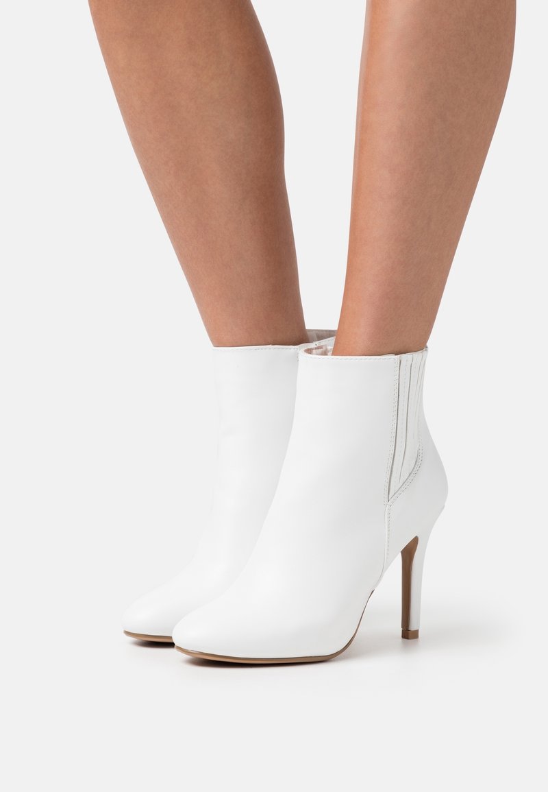 Bottes blanches à talons hauts jusqu'à la cheville portées sur des jambes nues avec des bouts pointus et des talons fins sur fond blanc.