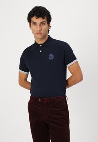 Mornarice plava polo majica s sivim manžetama i izvezenim grbom. Uparen s bordo ženskim pamučnim hlačama, model stoji naslonjen na bijelu pozadinu.