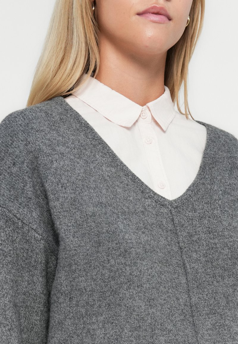Pull en laine gris avec un col en V, porté sur une chemise à col rose clair ; caractérisé par une texture douce et une coupe décontractée.