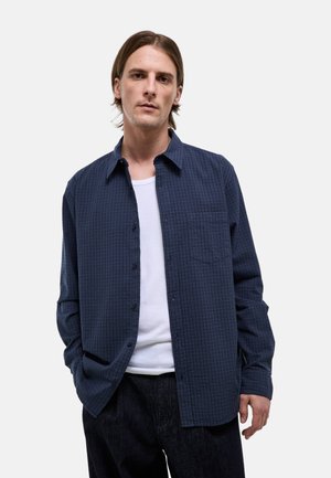 Jeune homme aux cheveux bruns mi-longs portant une chemise à carreaux bleu marine boutonée sur un t-shirt blanc, debout avec une main dans la poche.