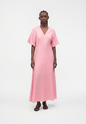Femme aux cheveux tressés portant une longue robe rose texturée avec des manches volantées et des sandales ouvertes marron, debout devant un fond blanc.