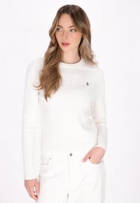 Maglione bianco a coste con girocollo e maniche lunghe, che presenta un piccolo emblema di un'ancora nera sul petto. Indossato con pantaloni bianchi.
