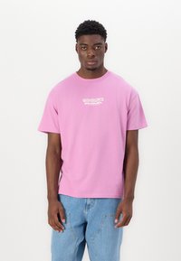 T-shirt en coton rose à manches courtes avec un col rond et un logo graphique blanc sur la poitrine. Associé à un jean en denim bleu clair.