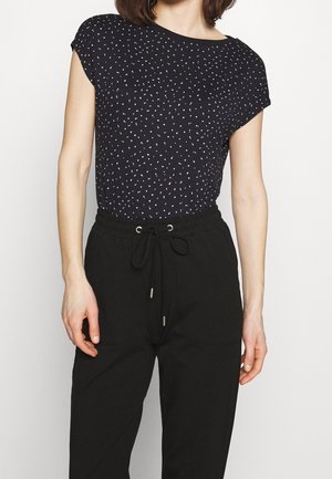 Vrouw draagt een zwart topje met korte mouwen en witte stippen, ingestopt in een zwarte, hooggetailleerde broek met trekkoord in de taille.