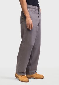 Homem com calças cinza largas com um pequeno bordado de cacto no bolso de trás e botas de trabalho bege, em fundo branco.