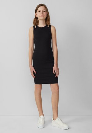 Schwarzes ärmelloses Kleid mit Schulterauschnitten, figurbetonter Schnitt, knielang. Kombiniert mit weißen Sneakers. Glatte Textur, einfache Silhouette.