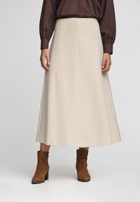 Kvinde iført en kremfarvet midi-nederdel, brun langærmet bluse og brune ruskindsankelstøvler, der står foran en ensfarvet baggrund.