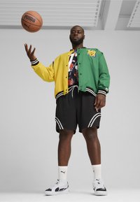 Farverig bomberjakke i gul og grøn, over en mønstret skjorte, parret med sorte shorts med hvide zigzag-accenter og sorte sneakers.