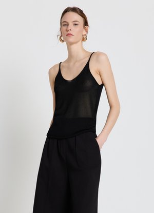 Jeune femme à la peau claire et cheveux attachés, portant des boucles d'oreilles créoles dorées, un haut noir sans manches et un pantalon noir ample, debout les mains dans les poches.