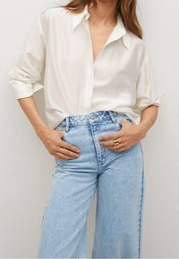 Femme portant une chemise blanche ample aux manches retroussées et un jean bleu clair taille haute, les mains posées sur les hanches avec une bague en or à la main droite.