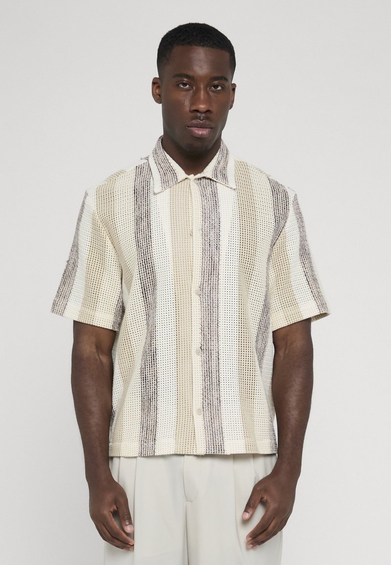 Homme portant une chemise à manches courtes boutonnée avec des rayures verticales beige, crème et grises, debout devant un fond clair uni.