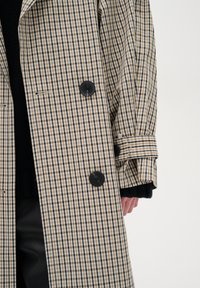 Cappotto trench doppiopetto a quadri beige e nero con grandi bottoni neri, polsini a contrasto e un design strutturato.