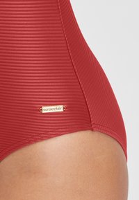Maillot de bain rouge à côtes avec un tissu texturé, présentant une petite étiquette dorée marquée "sunseeker." Met l'accent sur un gros plan du côté inférieur.