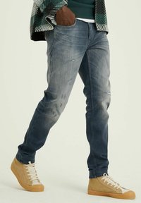 Džínové kalhoty ve vybledlém modrém odstínu s opotřebovanými detaily. Spárovány s hnědými vysokými teniskami se bílými tkaničkami a texturovanými látkovými akcenty.