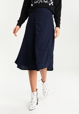 Kobieta w czarnym swetrze z białym kwiatowym haftem, granatowej midi spódnicy w wzory oraz białych botkach na obcasie z ćwiekami.