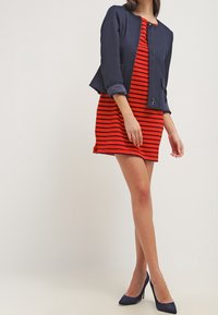 Veste courte bleu marine sur une robe rouge à rayures noires, associée à des talons hauts bleus. Le tissu semble lisse ; la veste possède une fermeture éclair.
