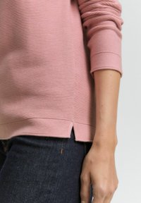 Close-up van de arm van een persoon die een roze geribbelde langemouwen top en donkerblauwe jeans met een kleine zijsplit aan de bovenkant draagt.