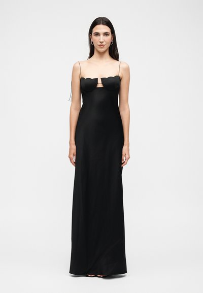 Femme aux longs cheveux foncés portant une robe noire longue jusqu'au sol avec de fines bretelles et un détail festonné au niveau de la poitrine, debout sur un fond blanc uni.