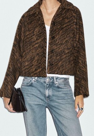 Person iført en brun mønstret cropped knapjakke, hvid top, lyseblå jeans, der holder en mørkebrun håndtaske.