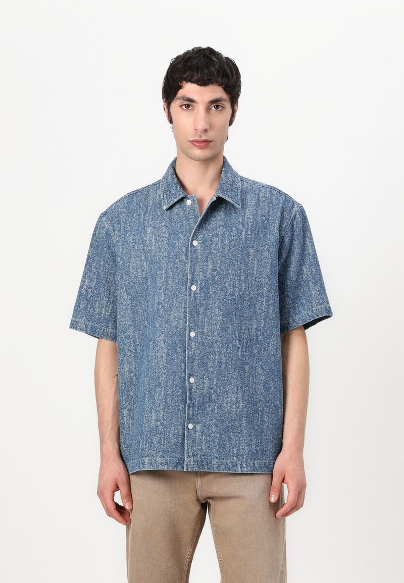 Camicia a maniche corte in denim azzurro chiaro con un motivo testurizzato, apertura frontale a bottoni e colletto classico. Dettaglio di cuciture bianche sottili.