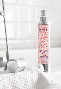 Rensegel i en transparent flaske med lyserøde perler, sølvpumpe og blå tekst. Ved siden af er der et skinnende sølvhætte på et struktureret hvidt håndklæde.