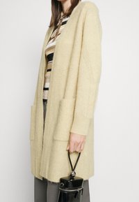 Ljusgul, lång kofta gjord av fluffigt material, med öppen framsida, två framfickor och ribbade ärmslut. Håller en liten svart väska.