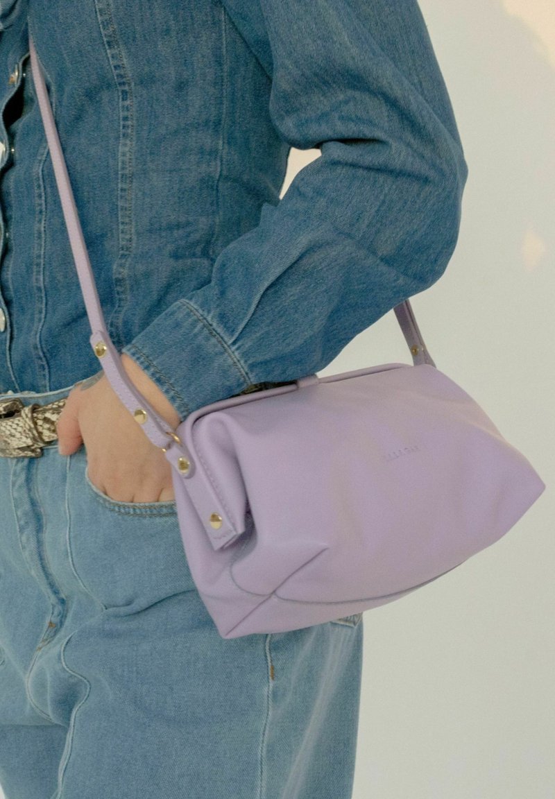 Bolso de mano de cuero lavanda con un diseño fruncido, herrajes en tono dorado y una correa delgada y ajustable, sostenido contra un outfit de denim.