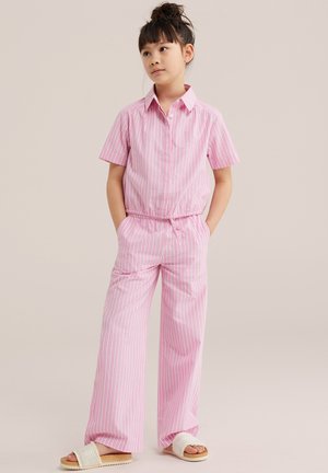 Mladé dievča so zaviazanými tmavými vlasmi, oblečené v ružovo-bielom pruhovanom tričku s krátkymi rukávmi a zodpovedajúcich širokých nohaviciach, na nohách béžové sandále.