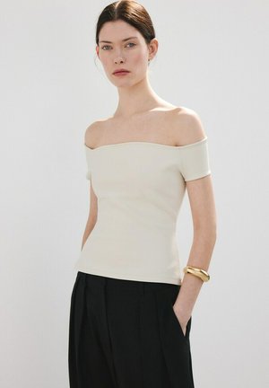 Femeie purtând un top crem cu umerii goi și pantaloni negri, stând cu o mână în buzunar și o brățară aurie la încheietură, pe un fundal neutru.