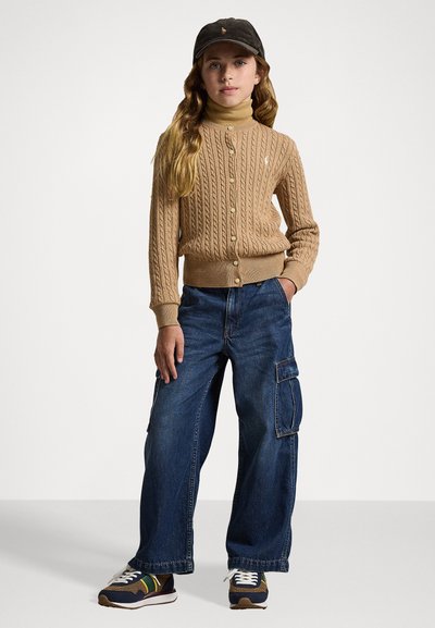 Μπεζ πλεκτή ζακέτα με πέντε κουμπιά, φορεμένη πάνω από ένα ανοιχτόχρωμο τζάκετ. Μπλε φαρδιά τζιν με τσέπη cargo. Σκούρος καπέλο και αθλητικά παπούτσια με πολύχρωμες λεπτομέρειες.