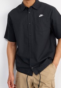 Černá krátká košile na knoflíky s kapsou na prsou, na které je bílé logo Nike. Má stahovací lem a hladkou texturu.