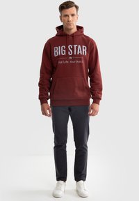 Bordeauxfarbener Kapuzenpullover mit "BIG STAR"-Aufdruck und Kängurutasche, kombiniert mit dunklen Hosen und weißen Turnschuhen.