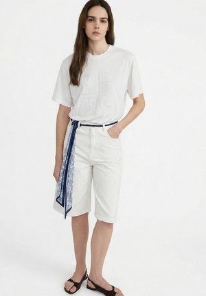 Vrouw met lang bruin haar die een wit T-shirt met reliëf draagt, witte knielange shorts, een marineblauwe riem met een patroon sjaal en zwarte open-toe schoenen.