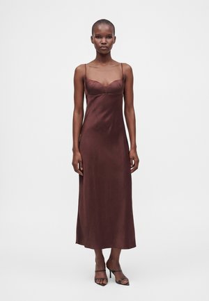 Femme portant une longue nuisette en satin brun foncé à fines bretelles et des escarpins noirs ouverts à l'avant, debout devant un fond blanc uni.