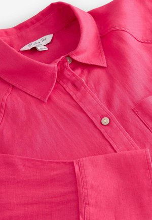 Camisa de lino rosa con cuello tipo polo, cierre de botones y bolsillo delantero. Presenta una textura suave y un sutil brillo.