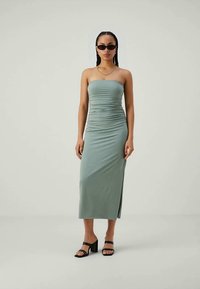Vestuário midi verde-claro, sem alças, com textura franzida e fenda lateral, combinado com óculos de sol pretos e sandálias de salto bloco.