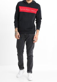 Sudadera azul marino con una franja horizontal roja que presenta el logo "Calvin Klein Jeans"; combinada con jeans oscuros y zapatillas blancas. Estilo casual.