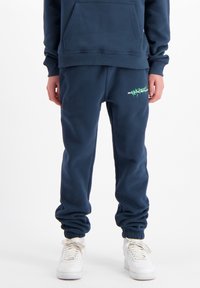 Navyblauwe sweatpants met elastische boorden, voorzien van een groene grafische tekening op de dij en zijzakken. Gecombineerd met witte sneakers.